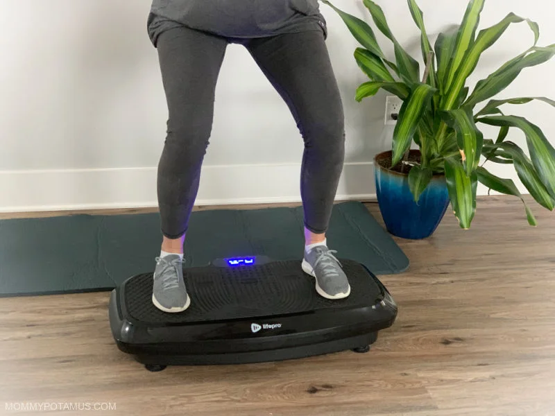 A person using a vibration therapy machine, standing on a platform with hands resting on the side handles.