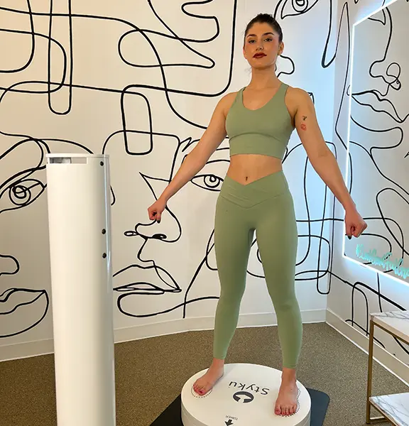 A young woman stands on a Styku body scanner, preparing for a comprehensive 3D body analysis.